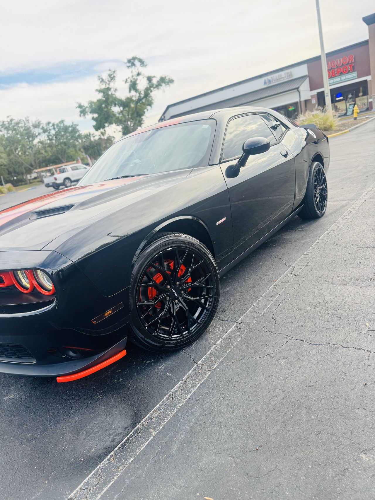 Black Dodge Challenger with glossy, freshly cleaned paint and red brake calipers parked on a paved lot in front of a liquor store and other businesses. The car wash result by Speed Shine is showcased through the mirror-like shine and spotless finish. The environment is urban with an overcast sky, creating an energetic and bold atmosphere that highlights the car's sporty and powerful presence.
