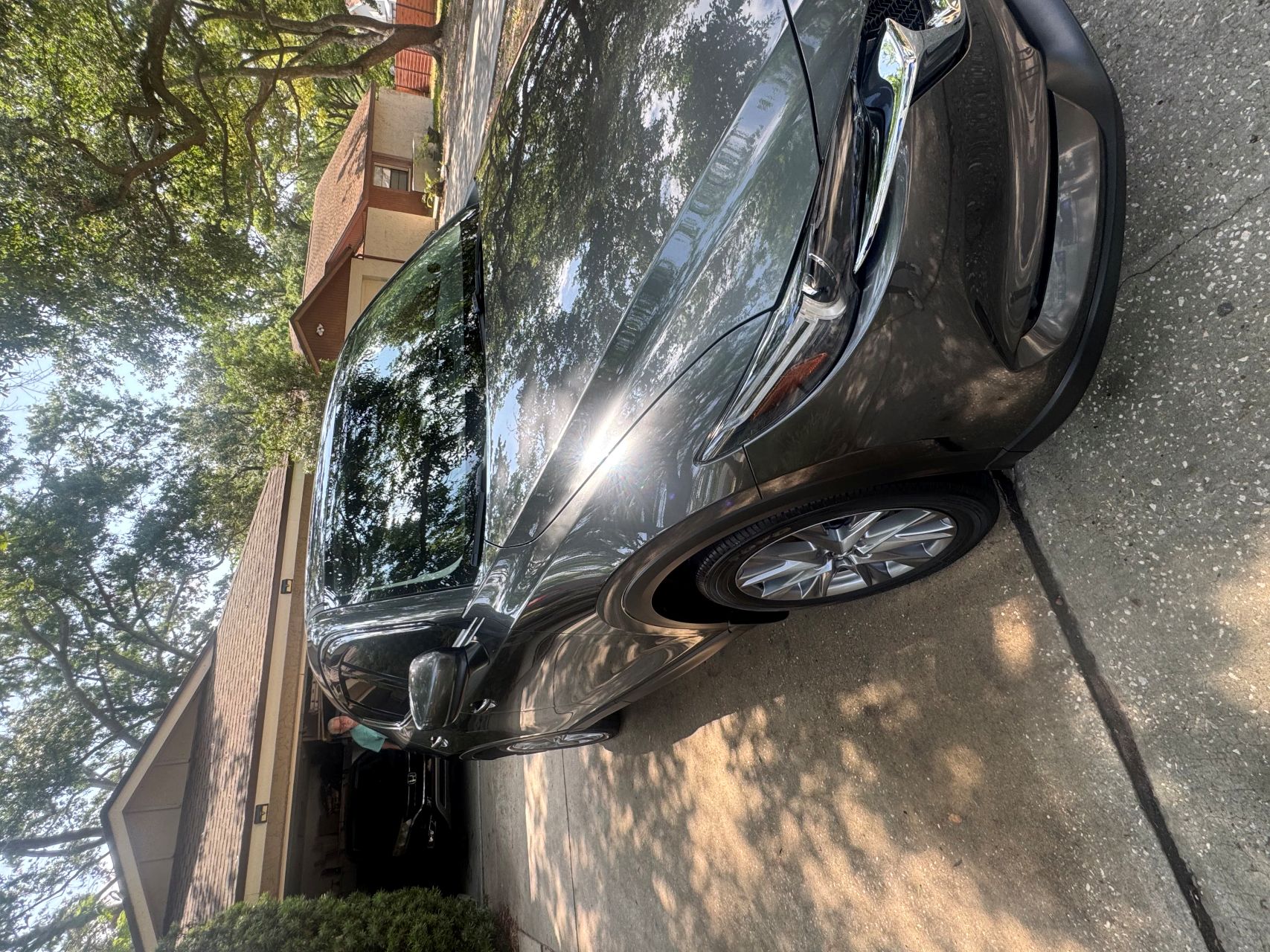 Gray Mazda CX-5 SUV parked on a driveway in front of a suburban house with trees casting shadows on the car. The vehicle has a clean, glossy finish and sunlight reflects off the hood and windshield.